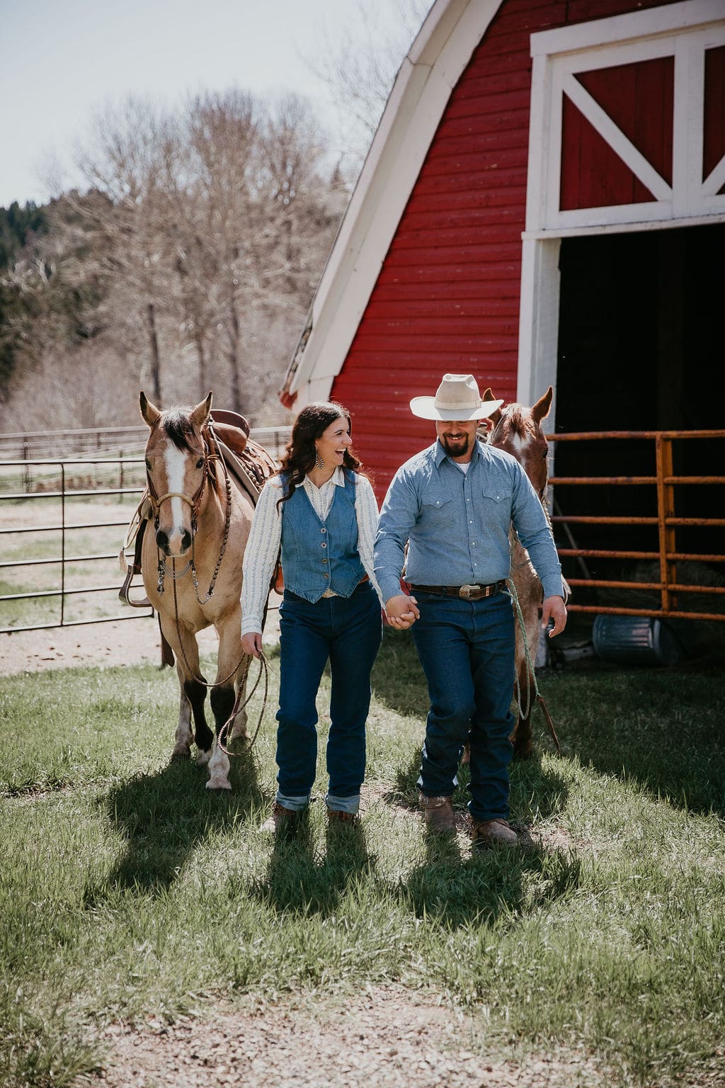 Engaged couple walking hand in hand with horses in front of a red barn during their Clyde Park, Montana ranch engagement session