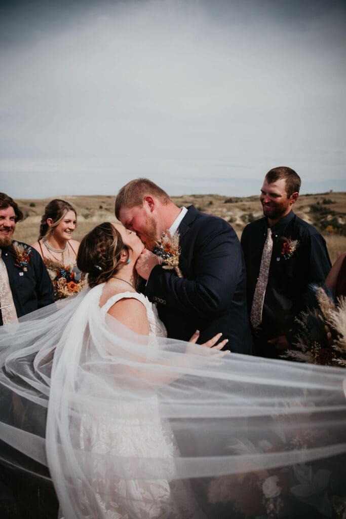 Bride and groom share kiss with flowing veil during ceremony as bridal party watches at Belfield wedding