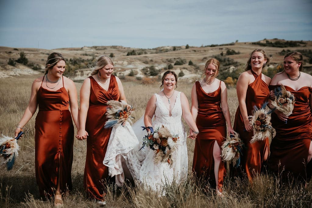 Bride walking through prairie fields with bridesmaids in rust-colored gowns during Belfield wedding celebration